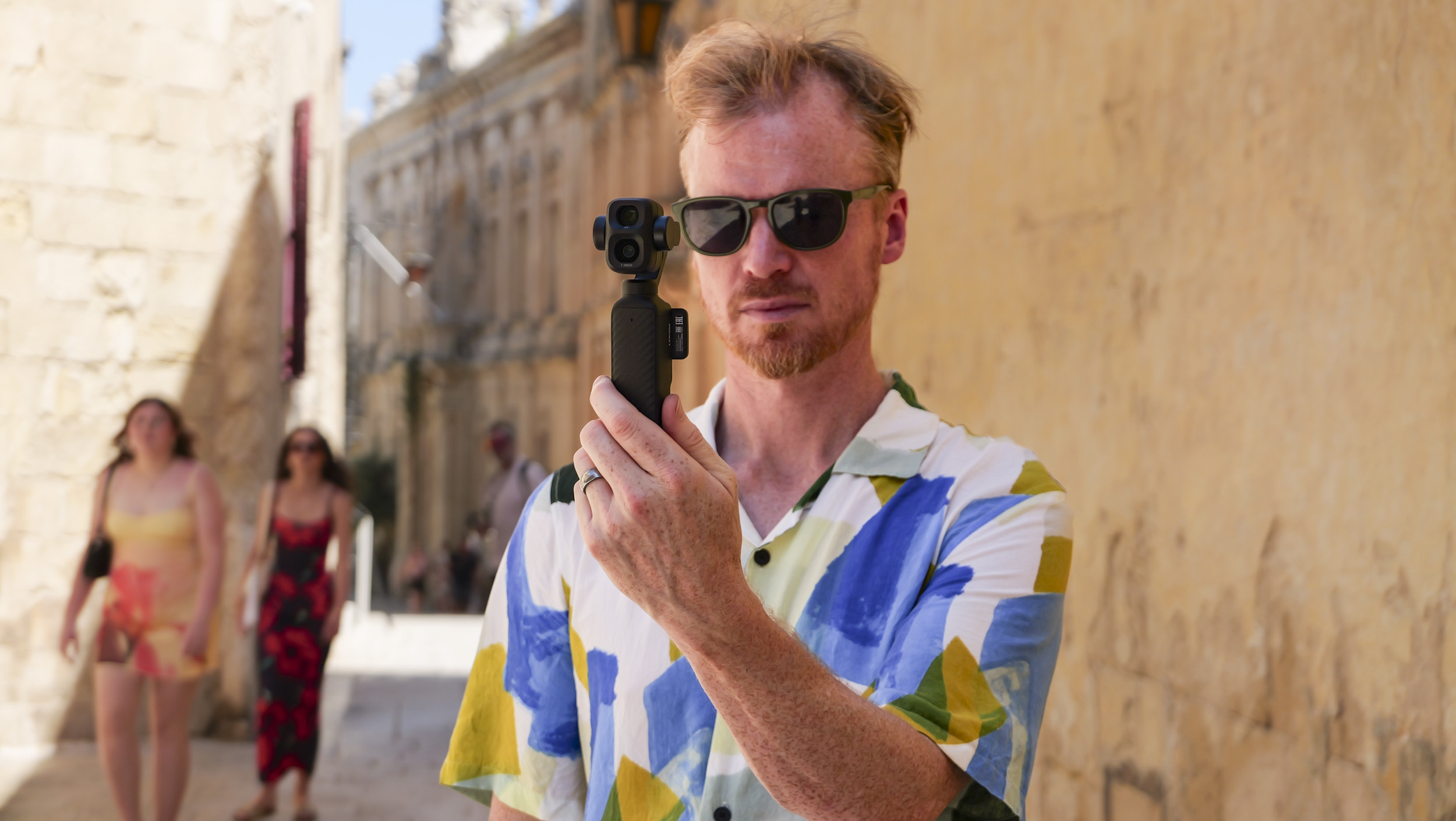 Man in bright summer short and shades, in an old town on a sunny day, holding the DJI Osmo Pocket 4P vlogging camera