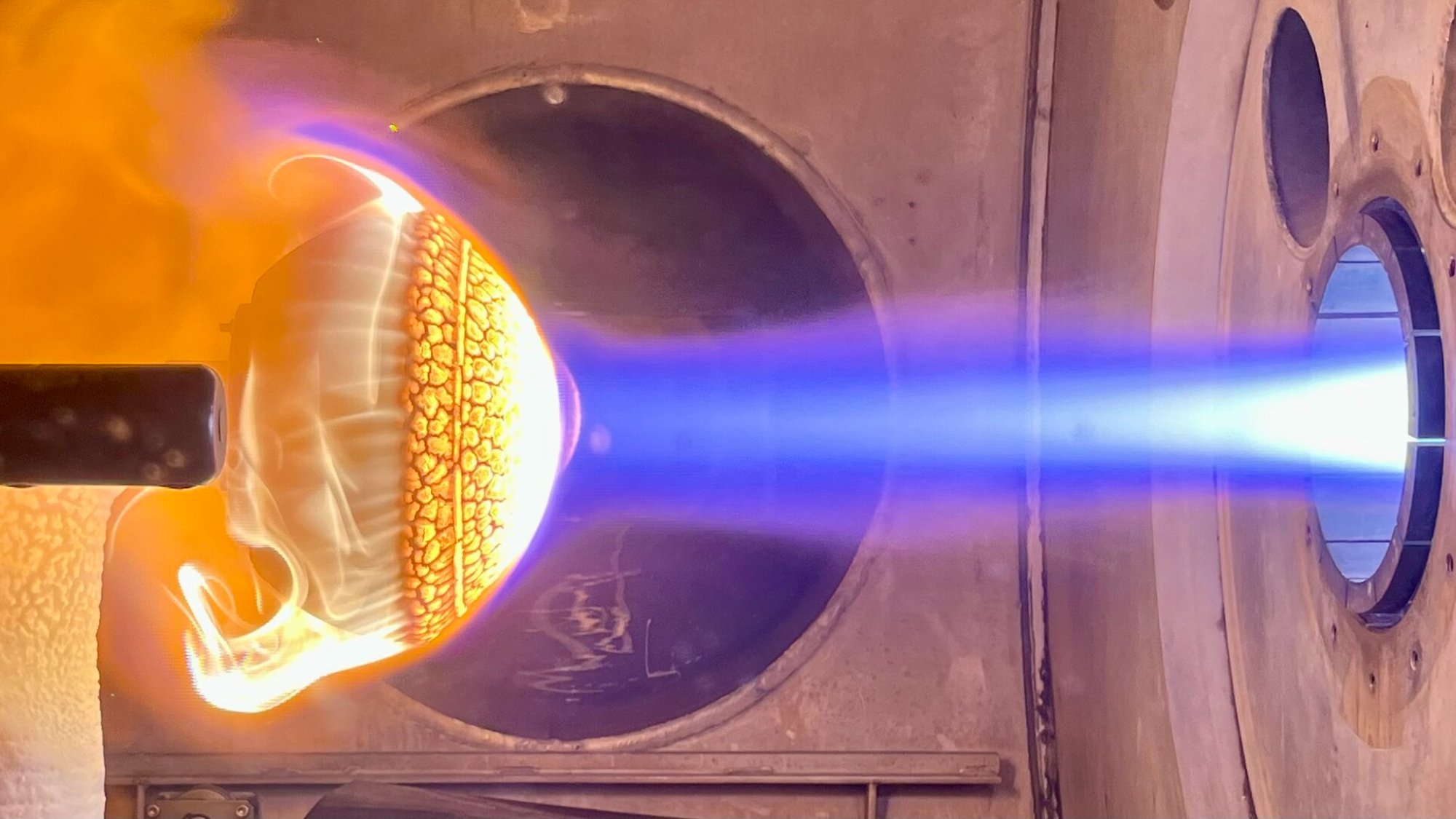 On the left is a heat shield that is bright orange and yellow engulfed in flames while on the right are bright blue and white flames pointed at the object.