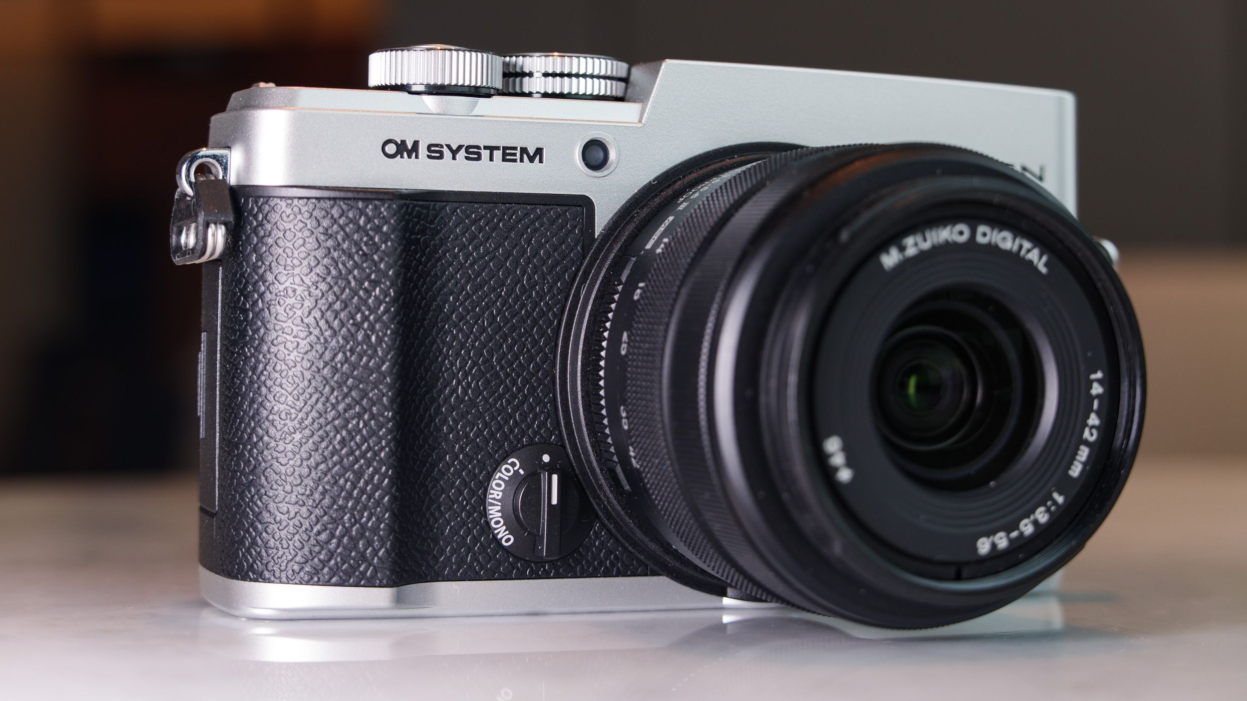 OM System PEN mirrorless camera with 14-42mm mark III kit lens attached, on a white marble surface in a modern room with lantern in the background