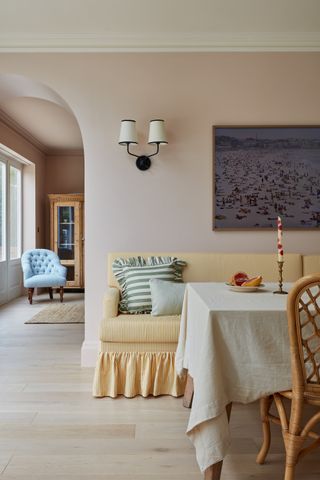 A light pink dining room with a curved archway that leads into a living room. There is a dining table with a white tablecloth and a yellow and white striped bench seat that has a ruffled skirt around the bottom. A plum painting hangs above the seat.