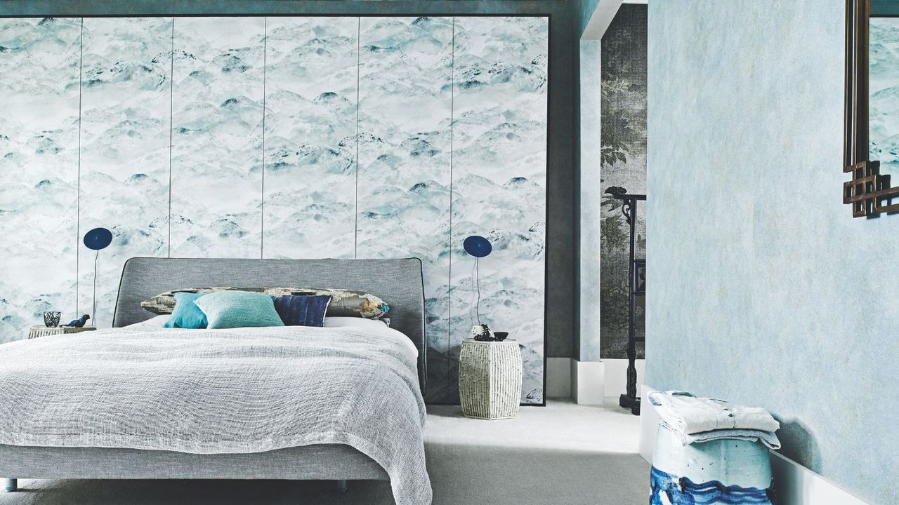 A blue bedroom with a grey double bed, statement wallpaper wall, two matching blue lamps and a blue patterned stool.