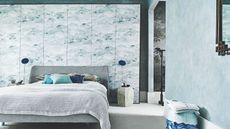 A blue bedroom with a grey double bed, statement wallpaper wall, two matching blue lamps and a blue patterned stool.