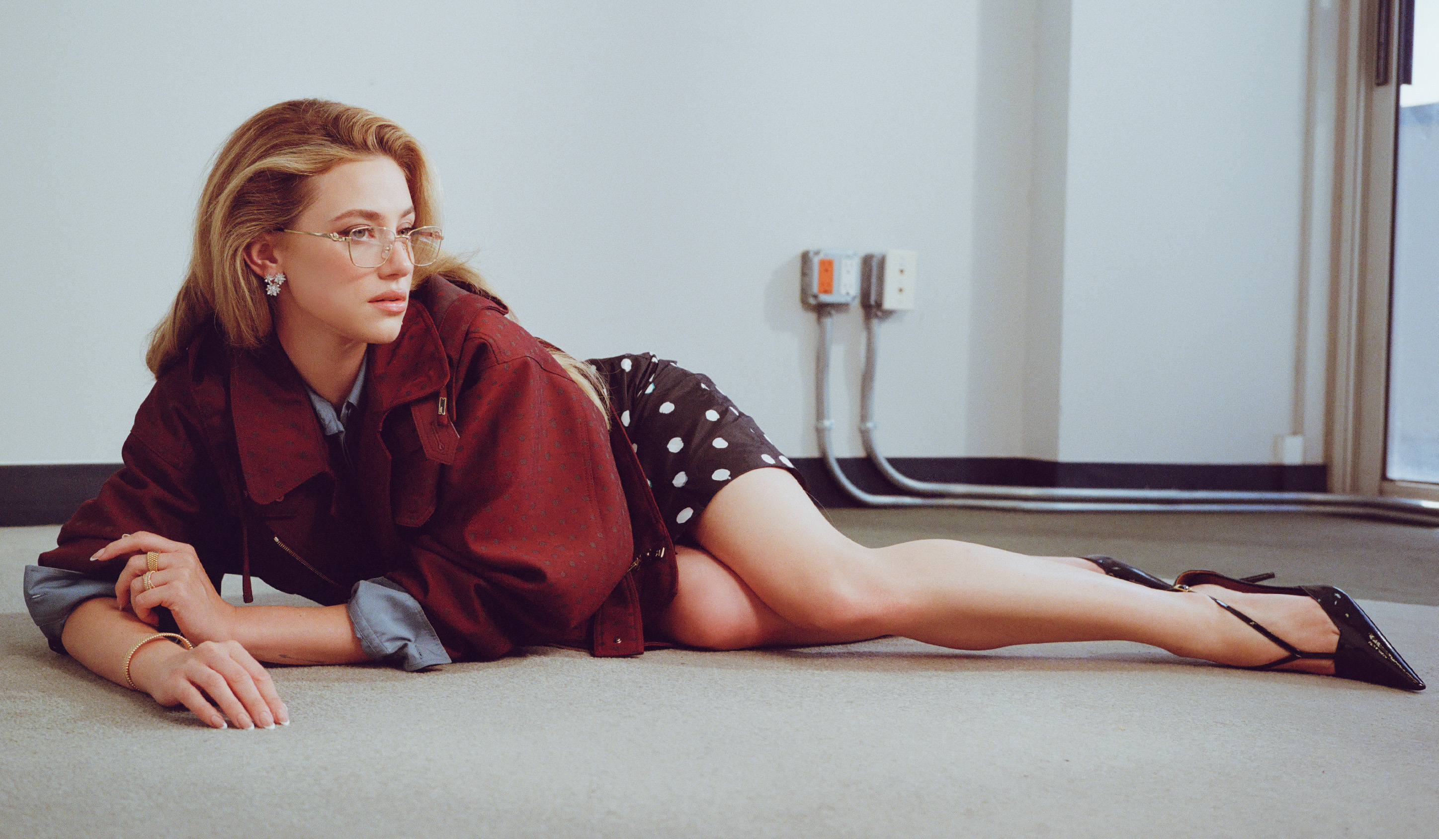 Lili Reinhart lying on the floor of an office building wearing clear glasses and a red jacket and polka dot skirt
