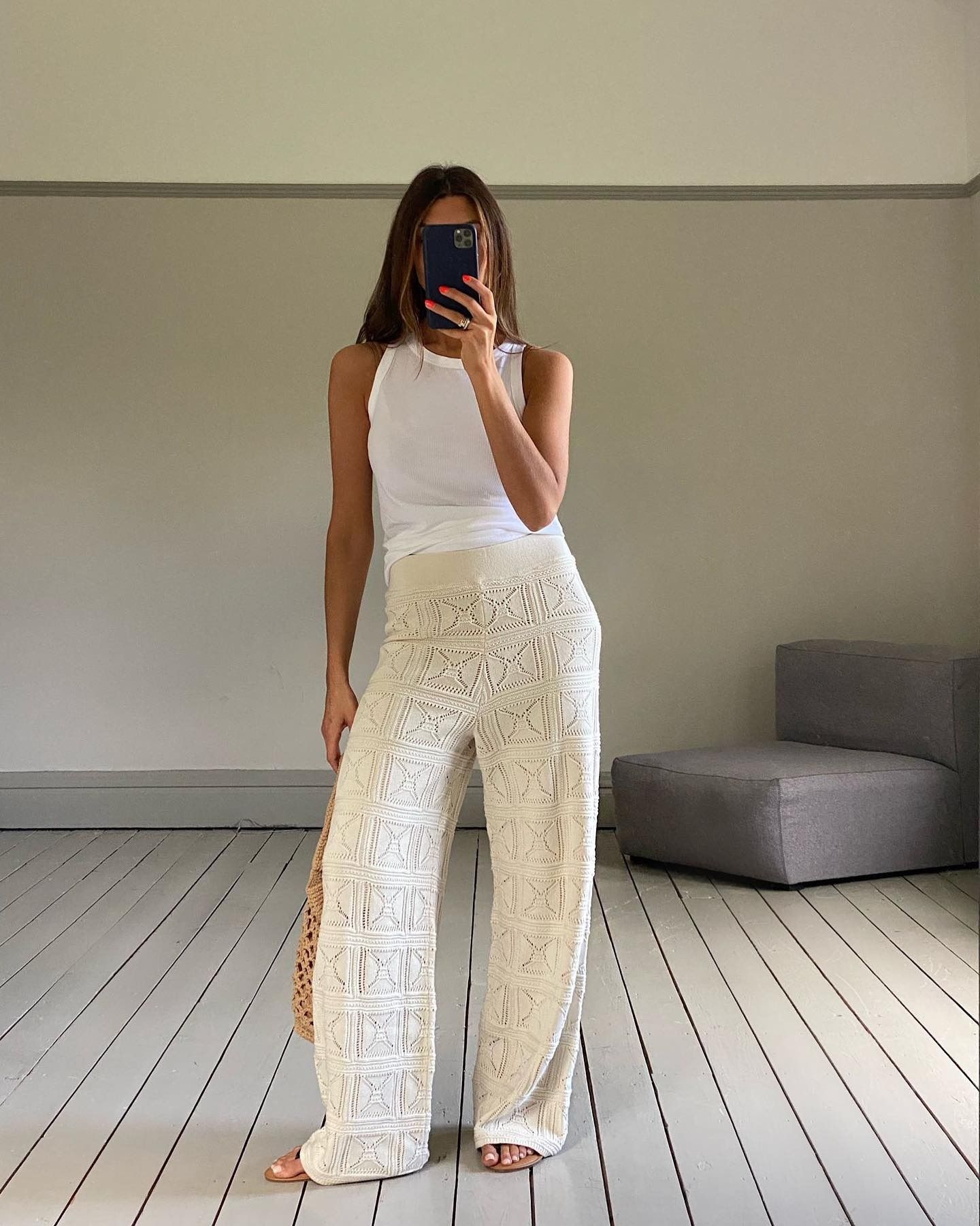 British influencer Marianne Smyth poses for a mirror selfie wearing a white tank top, ivory crochet wide-leg pants, a woven tote bag, and flat sandals