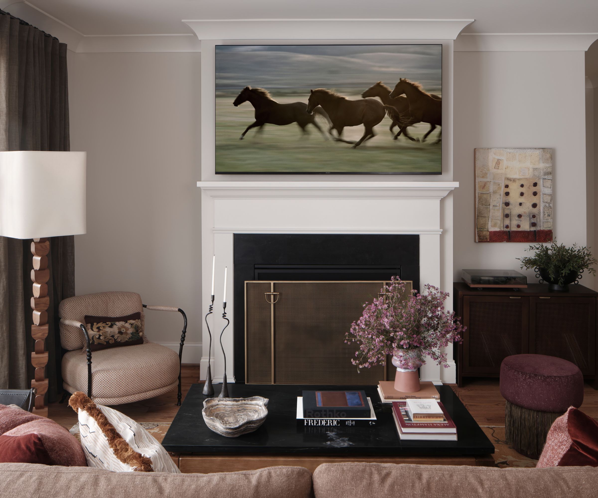 neutral living room with a white painted wooden fire surround, a large horse painting hung above the mantel with a taupe couch, small armchair facing the couch, and a purple small ottoman
