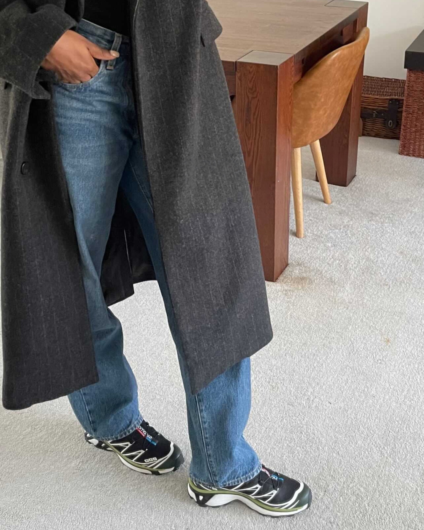 British influencer Marilyn Nwawulor-Kazemaks poses in her London home wearing a long gray pinstripe coat with straight-leg jeans and black Salomon sneakers.