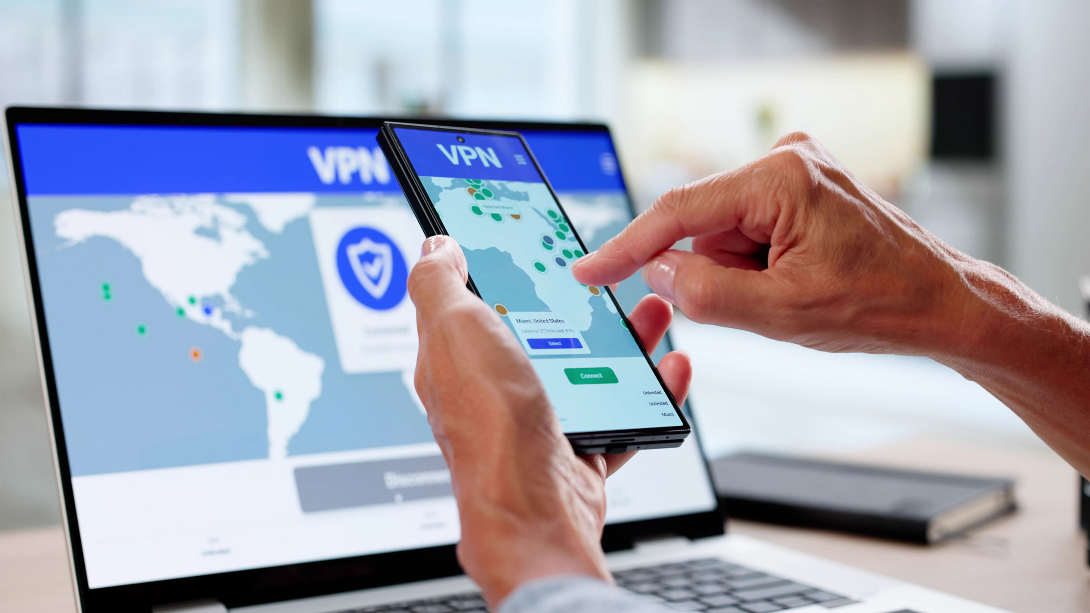 A hand is holding a mobile phone with a VPN on while a laptop with the same VPN screen is in the background, on a plain surface.