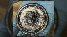 We see a circle-shaped silver disc with the face of medusa in the middle. A person wearing a blue glove is holding it.