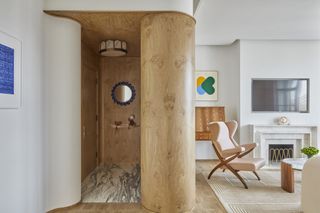 A whimsically decorated hotel suite juxtaposes warm wood surfaces, including a central wooden pillar, a coffee table, and a cabinet, with eccentric, colorful furniture such as a blue glass window, a pink shearling armchair, and striking prints.