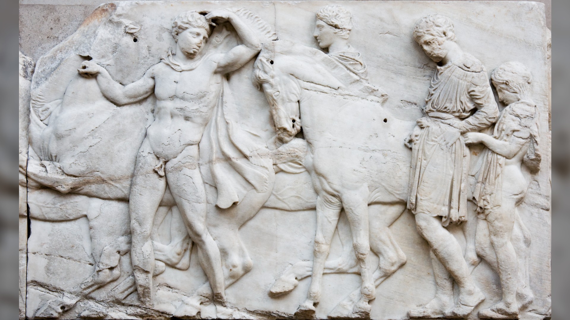 The Elgin Marbles stolen from Greece. From left to right we see a horse, a namke Greek man leading the way, whilst looking back at the others. Then there's another horse. Next to the horse is a person, and behind the horse if a person clothed in a tunic and a child with cloth draped around their shoulders.