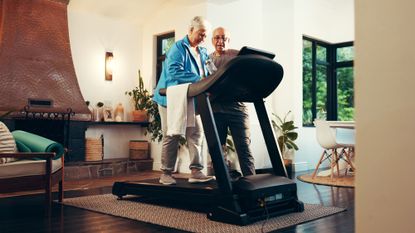 Woman walking on treadmill in domestic setting with man standing next to the treadmill and looking at the treadmill's console