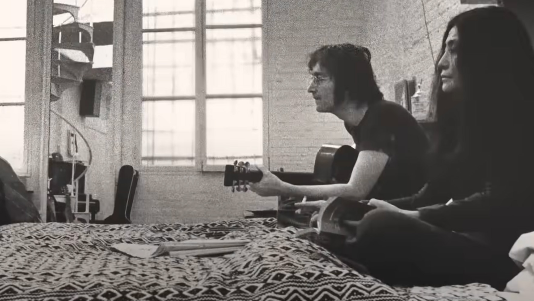 John Lennon and Yoko Ono, both sat on a bed, in One to One: John and Yoko.