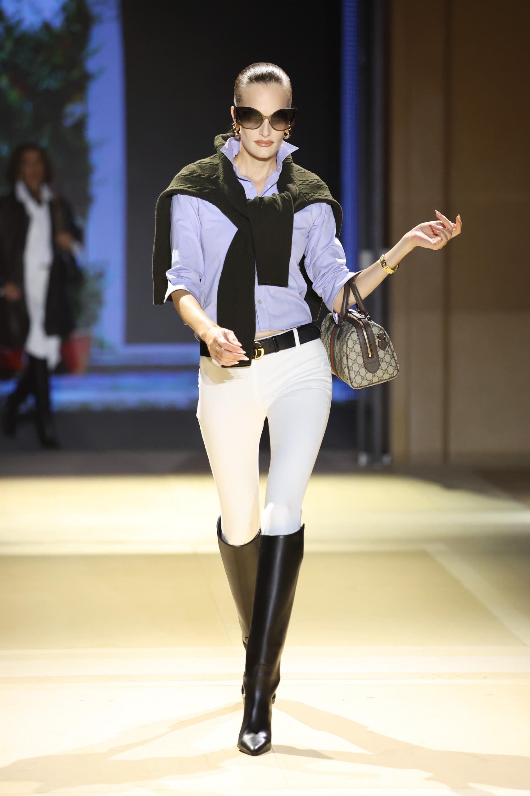 Model walks the Gucci SS27 Runway wearing jodhpur leggings tucked into a shirt with a jumper over the shoulder and black knee-high boots.
