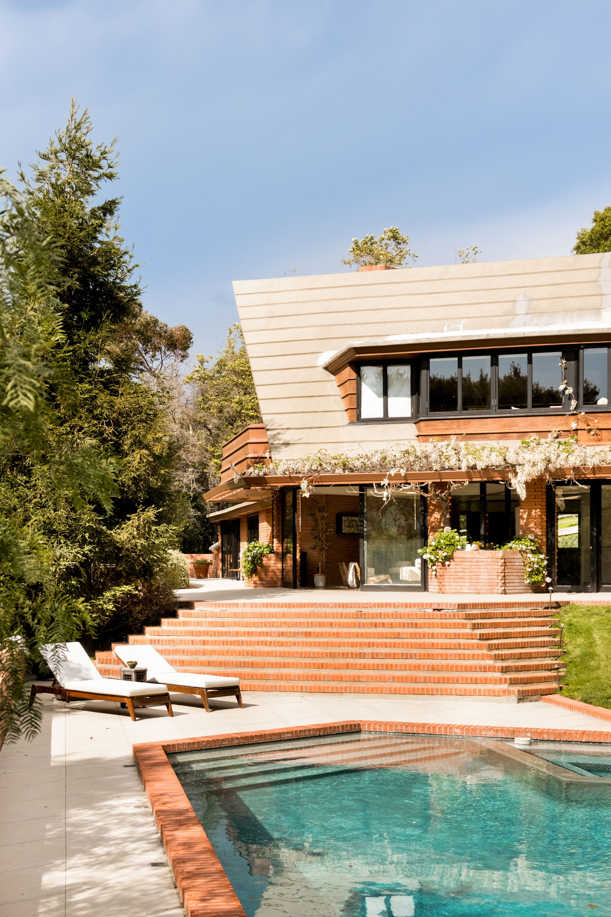 The exterior of a house with angular steps that lead to a pool area. The stairs and trim of the pool are made of traditional brick, while the house is wooden, and the pool deck is white. There are two lounge chairs by the pool.