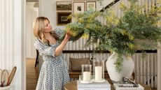 shea mcgee stood in a traditional entryway wearing a floral cottage style dress and preening a large vase of yellow flowers and foliage on a round wooden entryway table styled with coffee table books and pillar candles