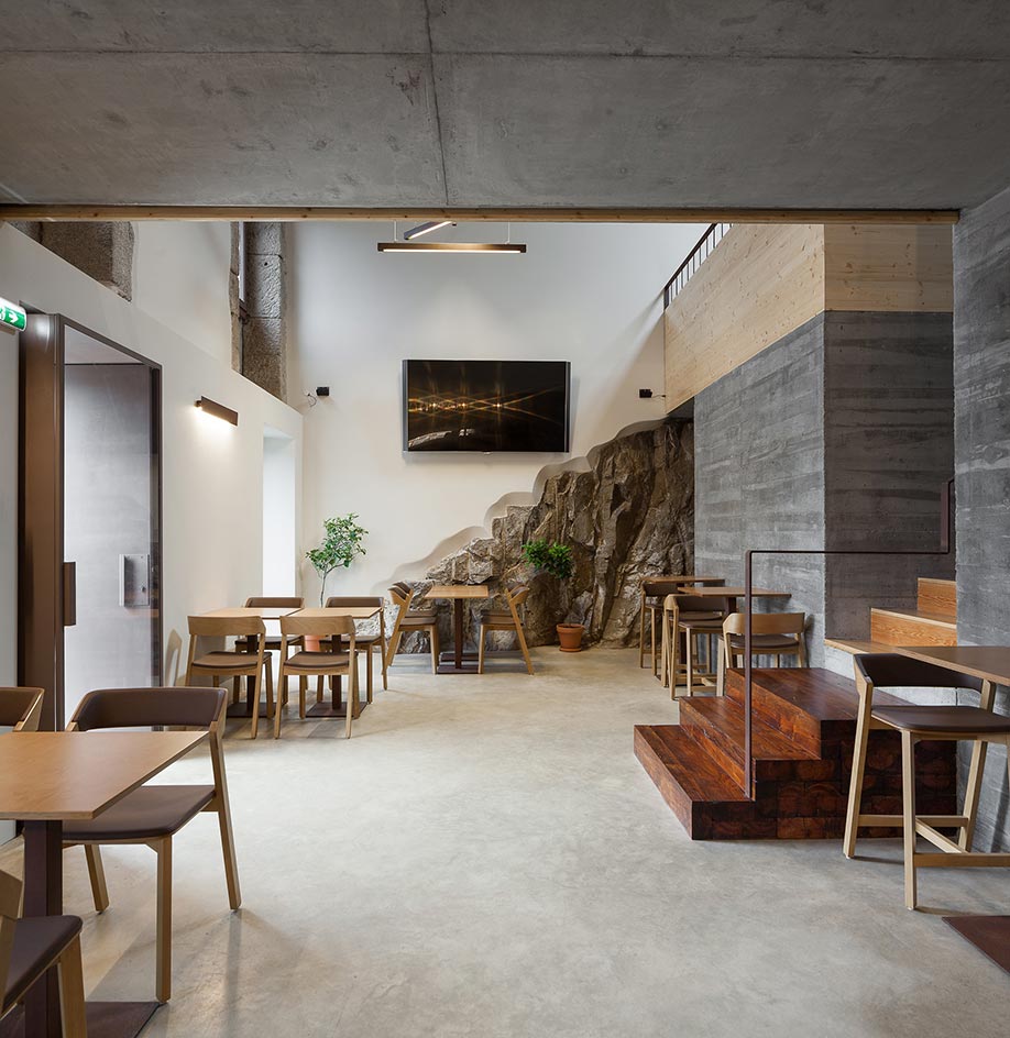 An interior view of the two-floor Entre Portas restaurant. The entrance is to the left. Floors, walls, and ceilings are all in gray concrete syle. The tables and chairs are all in light wood. To the right, there are wooden stairs that lead to the second floor.