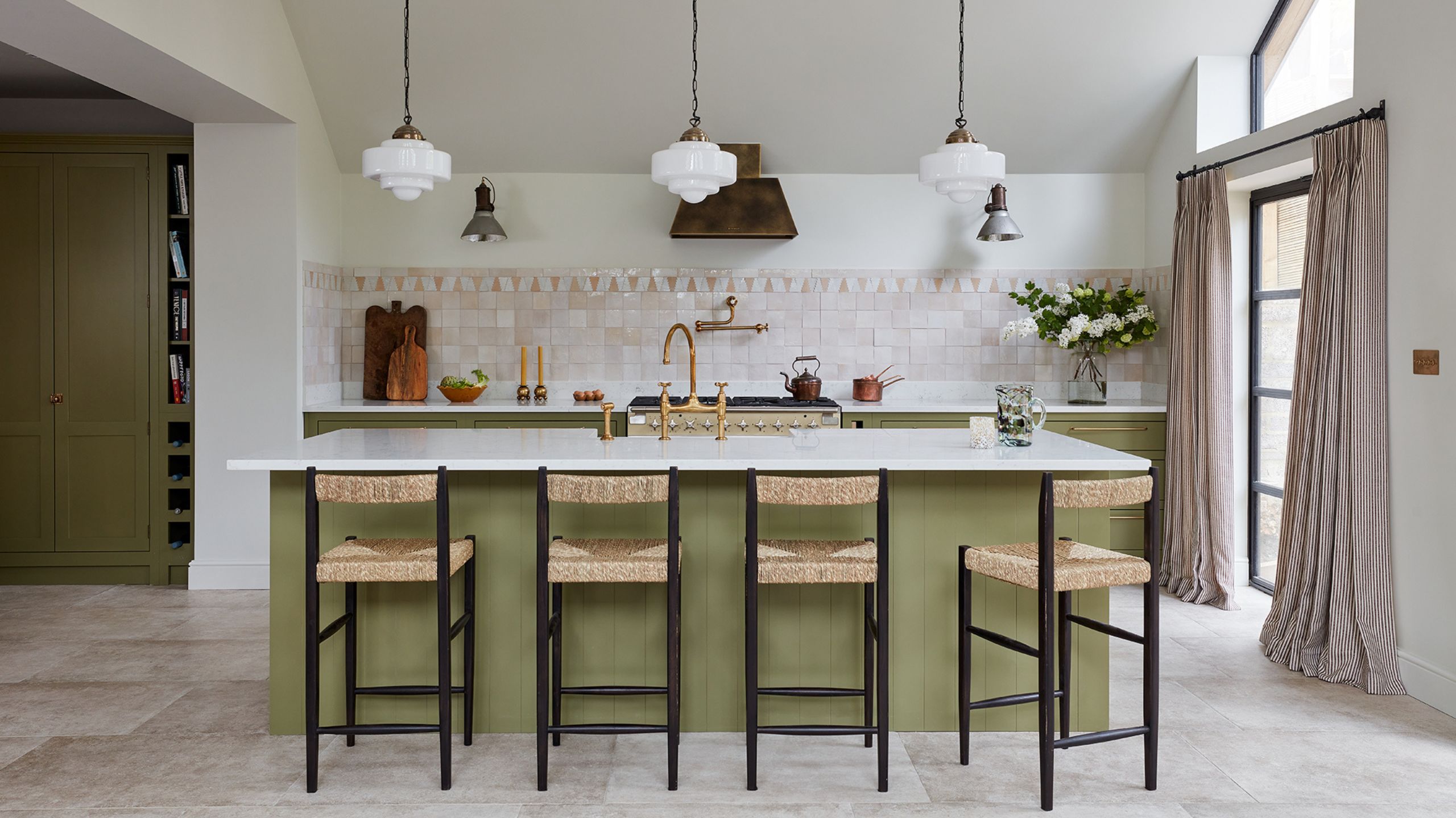Pink tiled kitchen with green island, rattan bar stools with black frames, and green cabinetry. To the right is a large crittal door with pink and white striped curtains.