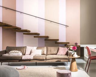 a living room with browns and pink paints as stripe on back wall, a wooden staircase, and a brown sofa with pink furniture and furnishings