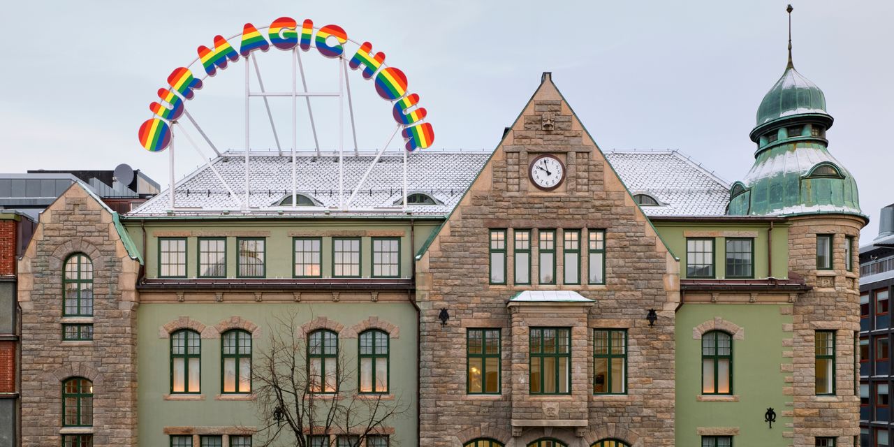 The brick and copper facade of a historic building is covered in a thin layer of snow at dusk and decorated with a rainbow-hued sign that reads &quot;OUR MAGIC HOUR&quot;