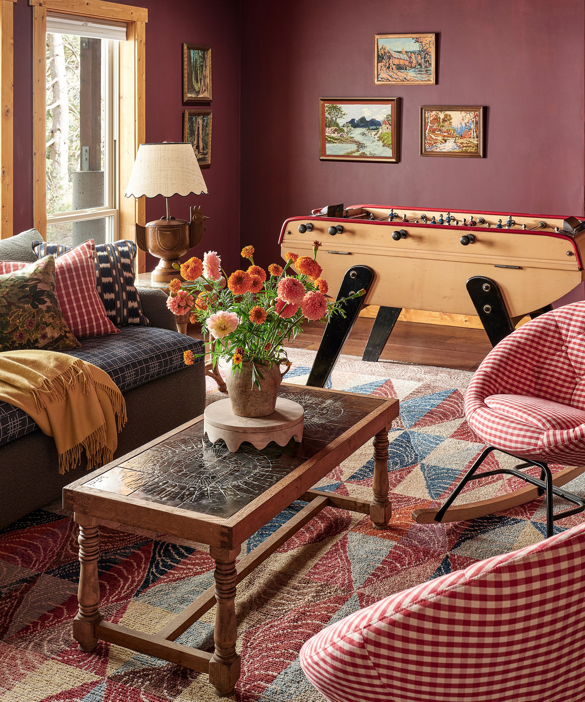 a moody dark red family room with a foosball table, red gingham chairs, folk rug and pine coffee table