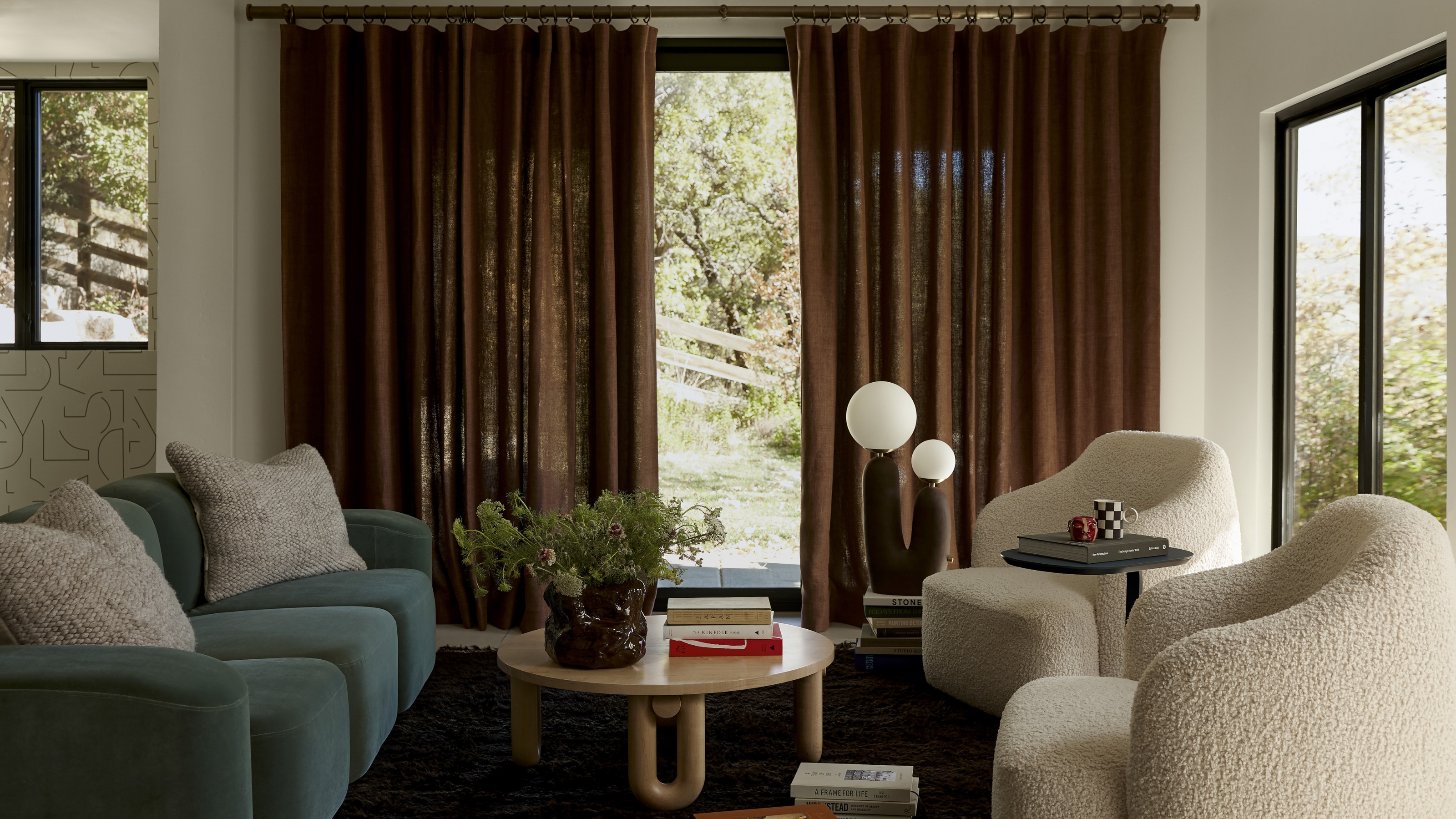 Image of a cozy living room with white walls and a dark rug. There are brown, semi-sheer curtains on the windows and two boucle, off-white accent chairs facing a teal sofa.