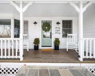 Styled farmhouse style porch decor with white rocking chairs, sage green front door, potted topiary trees, and greenery wreath.