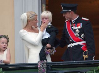 Crown Princess Mette-Marit in her wedding gown holding her son Marius and standing next to Crown Prince Haakon in a military uniform