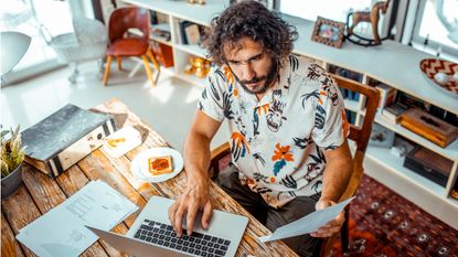 A casually dressed man works on a laptop at a table with breakfast nearby.