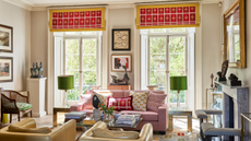 a living room with two yellow accent chairs, a pink couch with lots of pillows, two large windows with red shades, and lots of decor on the coffee table and throughout the room