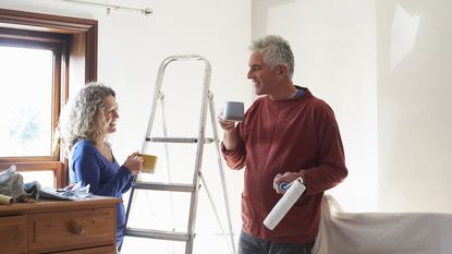 An older couple smile at each other as they drink coffee while taking a break from painting their bedroom.
