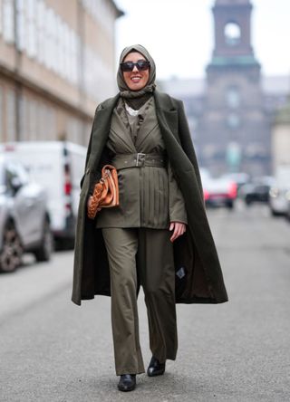 A guest wears a green khaki full outfit, sunglasses, a scarf over the head, a long coat, a waistcoat, a large belt, a brown leather bag, flared pants, leather pointed shoes, outside Lovechild 1979, during the Copenhagen Fashion Week AW24 on January 30, 2024 in Copenhagen, Denmark. (Photo by Edward Berthelot/Getty Images)