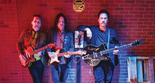 Texas Headhunters photographed with their guitars in front of. a red brick wall: [from left] Johnny Moeller, Ian Moore and Jesse Dayton