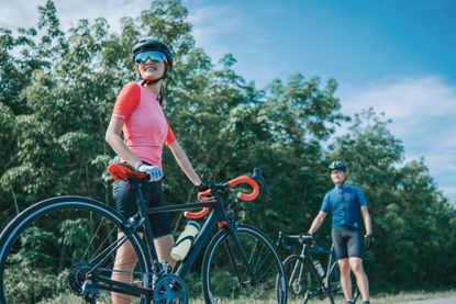 best bike accessories. This image shows a woman with her bike close to the camera, in the background in a man with his bike. They are standing on a road, there are trees in the background and blue sky. 