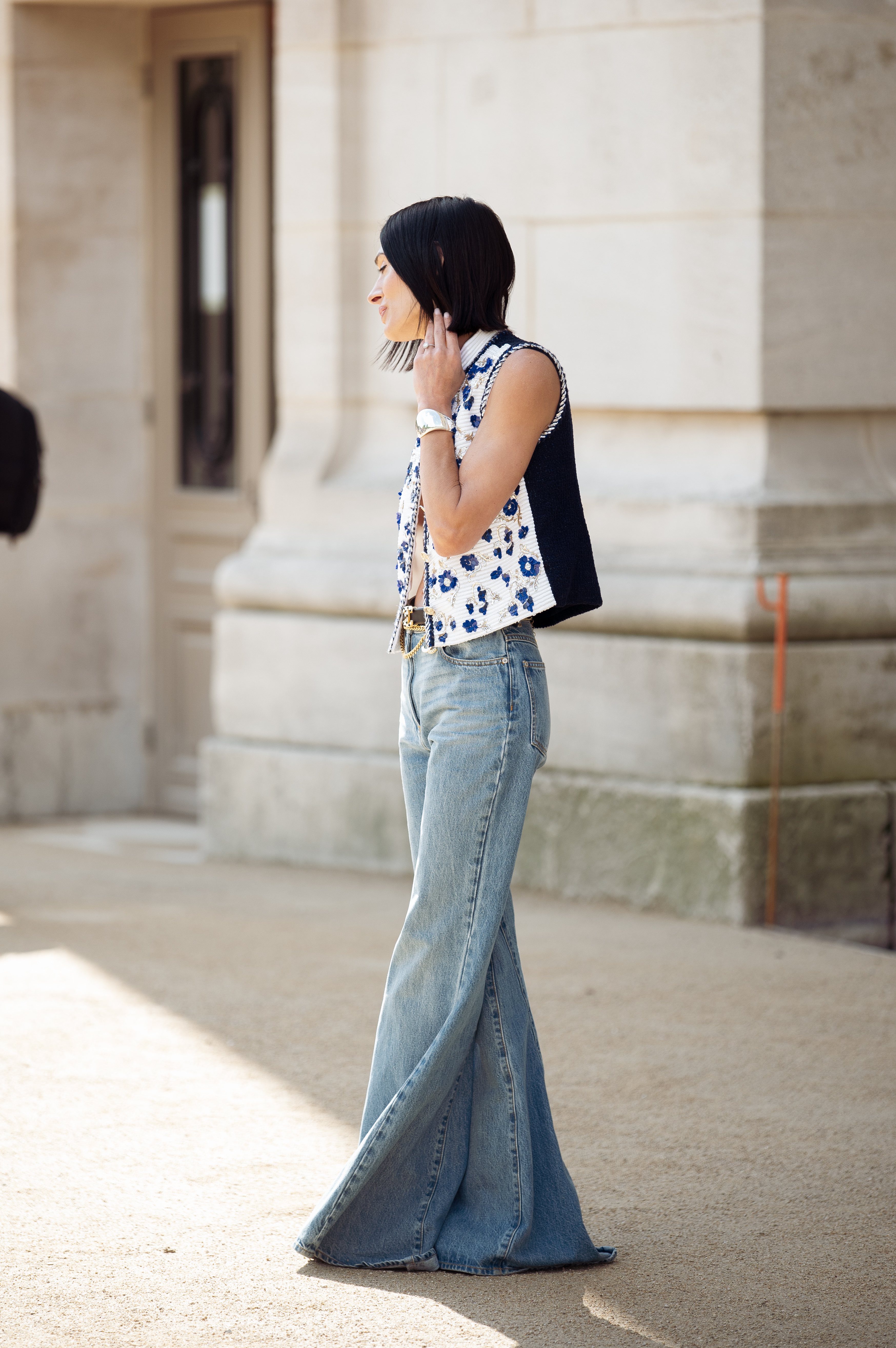 Una invitada viste jeans azules, chaleco blanco con adornos azules y dorados y cinturón de Chanel afuera del desfile de Chanel durante la Alta Costura Otoño/Invierno 2025/2026 como parte de la Semana de la Moda de París.