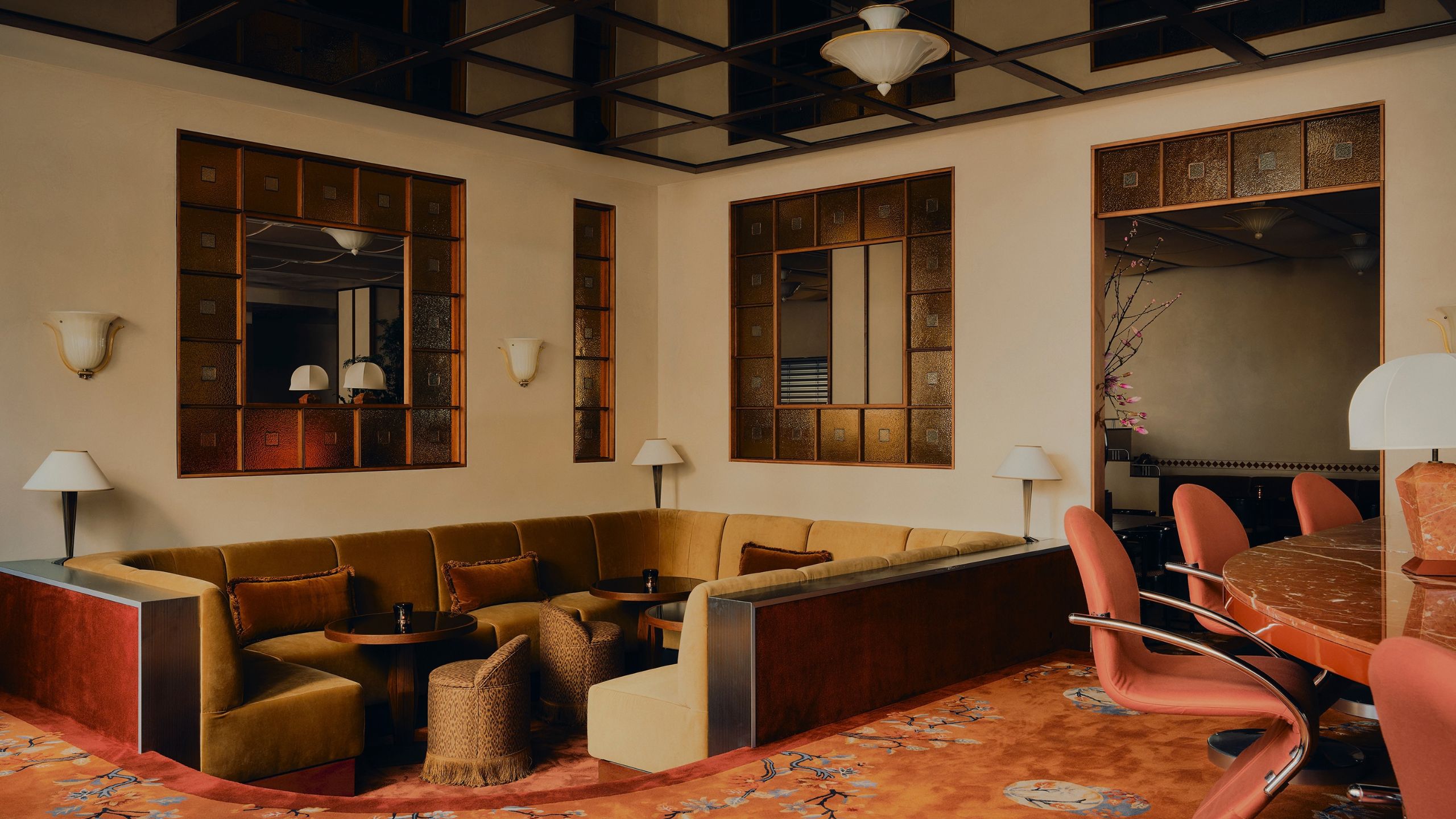 A modern conversation pit in a bar with stained glass windows, a mirror paneled ceiling and a floral carpet with trim, with ochre seating and brown throw pillows with small round bar tables and poufs