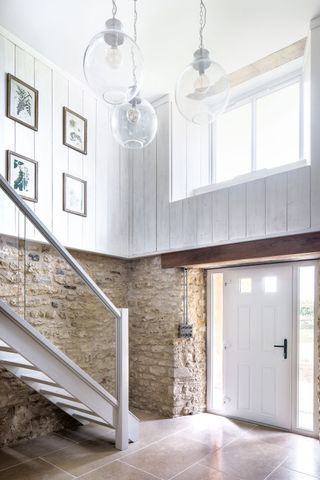 double height entrance hall with high window exposed stone lower half and painted wood paneling above three globe ceiling lights and glazed staircase