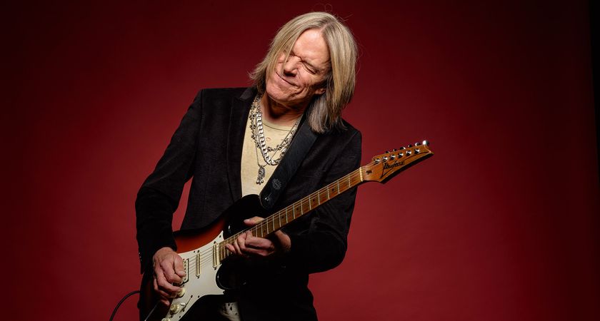 Andy Timmons bends a note and feels it as he plays his signature Ibanez electric against a deep red background. He wears a black jacket and a cream T-shirt and necklesses.