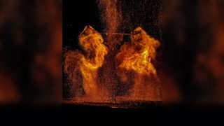 A night scene of a dramatic performance with molten iron or fire being thrown into the air, creating glowing orange explosions of sparks and flames shaped like wings against a black background