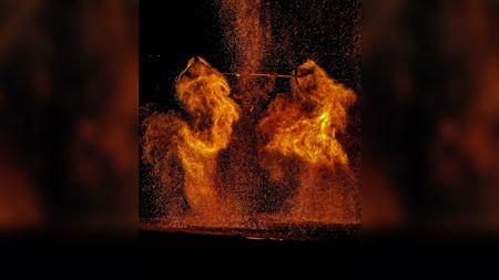 A night scene of a dramatic performance with molten iron or fire being thrown into the air, creating glowing orange explosions of sparks and flames shaped like wings against a black background