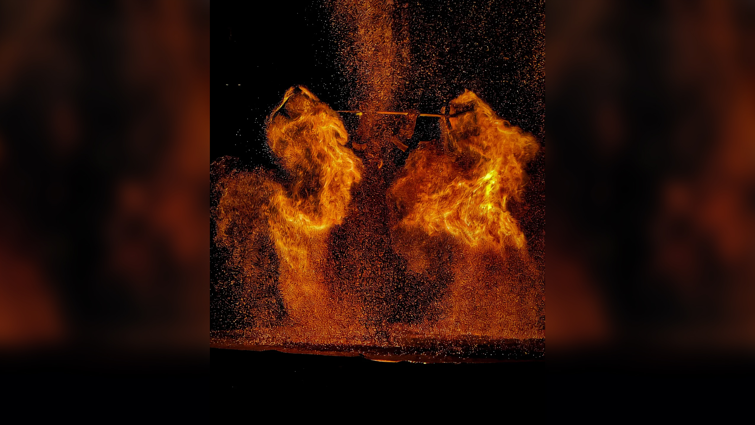 A night scene of a dramatic performance with molten iron or fire being thrown into the air, creating glowing orange explosions of sparks and flames shaped like wings against a black background