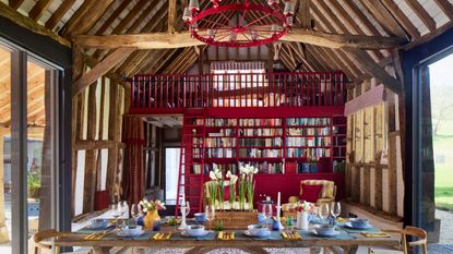 an oxfordshire barn with wooden beams, a vaulted ceiling, red chandelier, large wooden table and red painted library with a red ladder up to a mezzanine floor