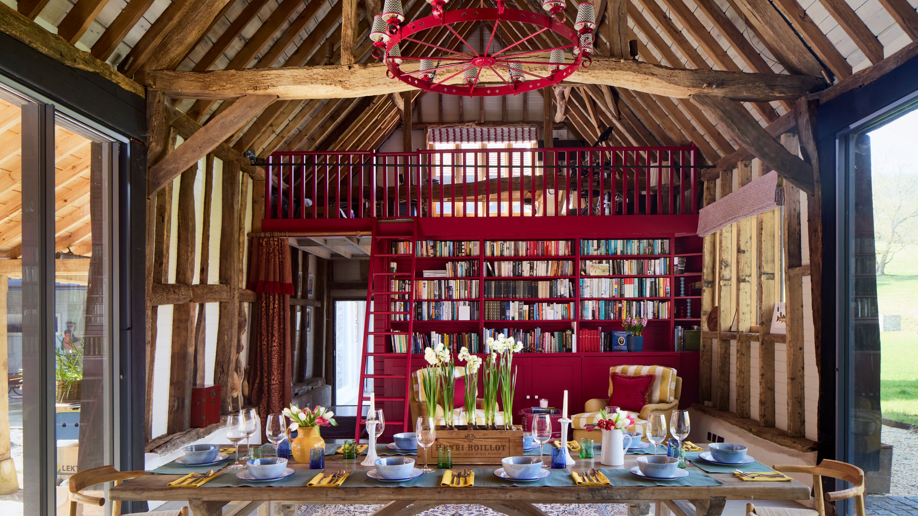 an oxfordshire barn with wooden beams, a vaulted ceiling, red chandelier, large wooden table and red painted library with a red ladder up to a mezzanine floor