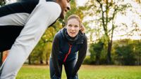 Photo of a man and woman outdoors, each standing with their hands on their knees as they recover from a workout. 