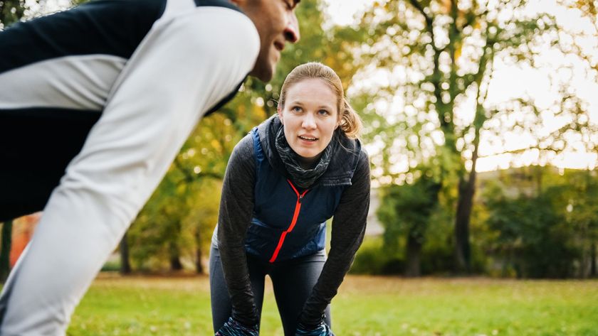 Photo of a man and woman outdoors, each standing with their hands on their knees as they recover from a workout. 