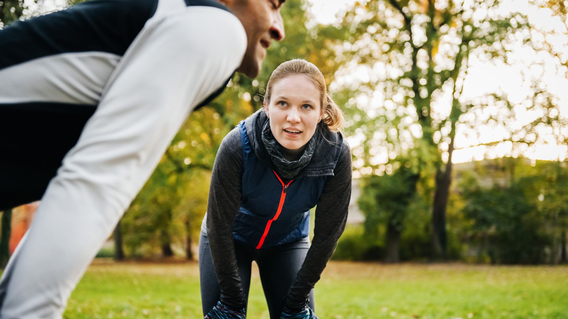 Photo of a man and woman outdoors, each standing with their hands on their knees as they recover from a workout. 