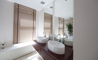 Double bathroom sink on dark wood surface with long mirror behind. Brown closed window blinds on left