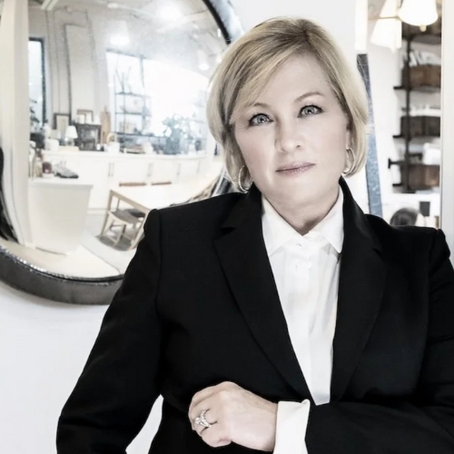 A woman standing in a black-and-white suit with a mirror behind her.