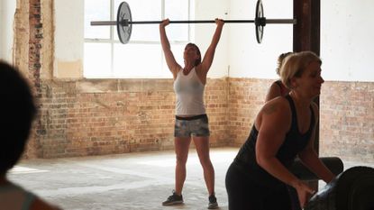 Woman lifting barbell with two plates above head in gym with other women lifting weights around