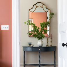 A hallway with a dark blue bobbin console table with a vase of leafy branches and a mirror hung above it