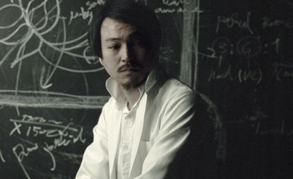 Makoto Azuma in a white shirt, sitting in front of a chalkboard